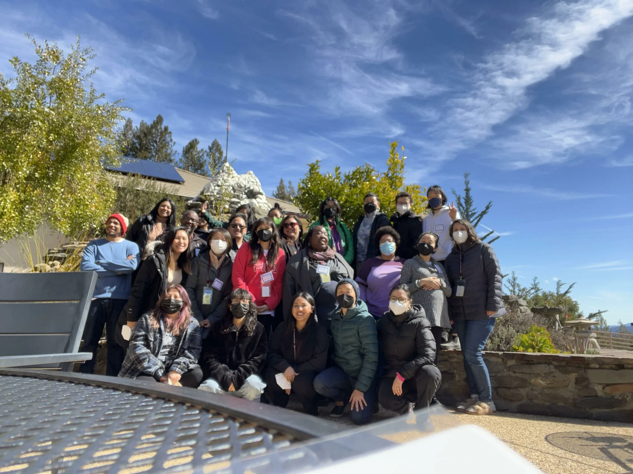 Group of diverse people wearing masks posing outdoors under a blue sky with clouds, surrounded by trees and plants.