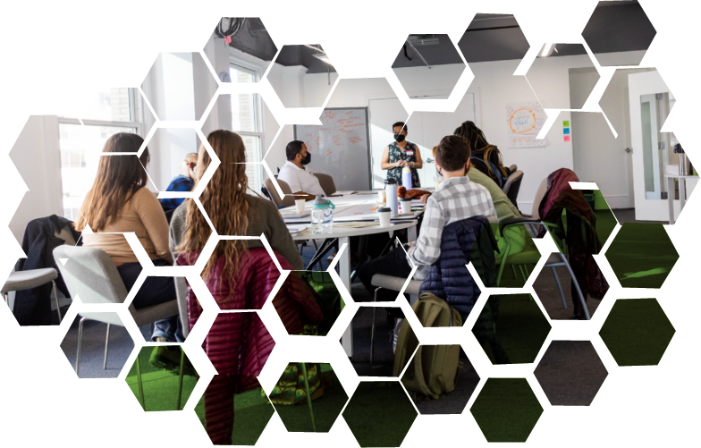 A group of diverse people attending a meeting in a bright conference room with large windows, a whiteboard, and a person standing and speaking at the front.