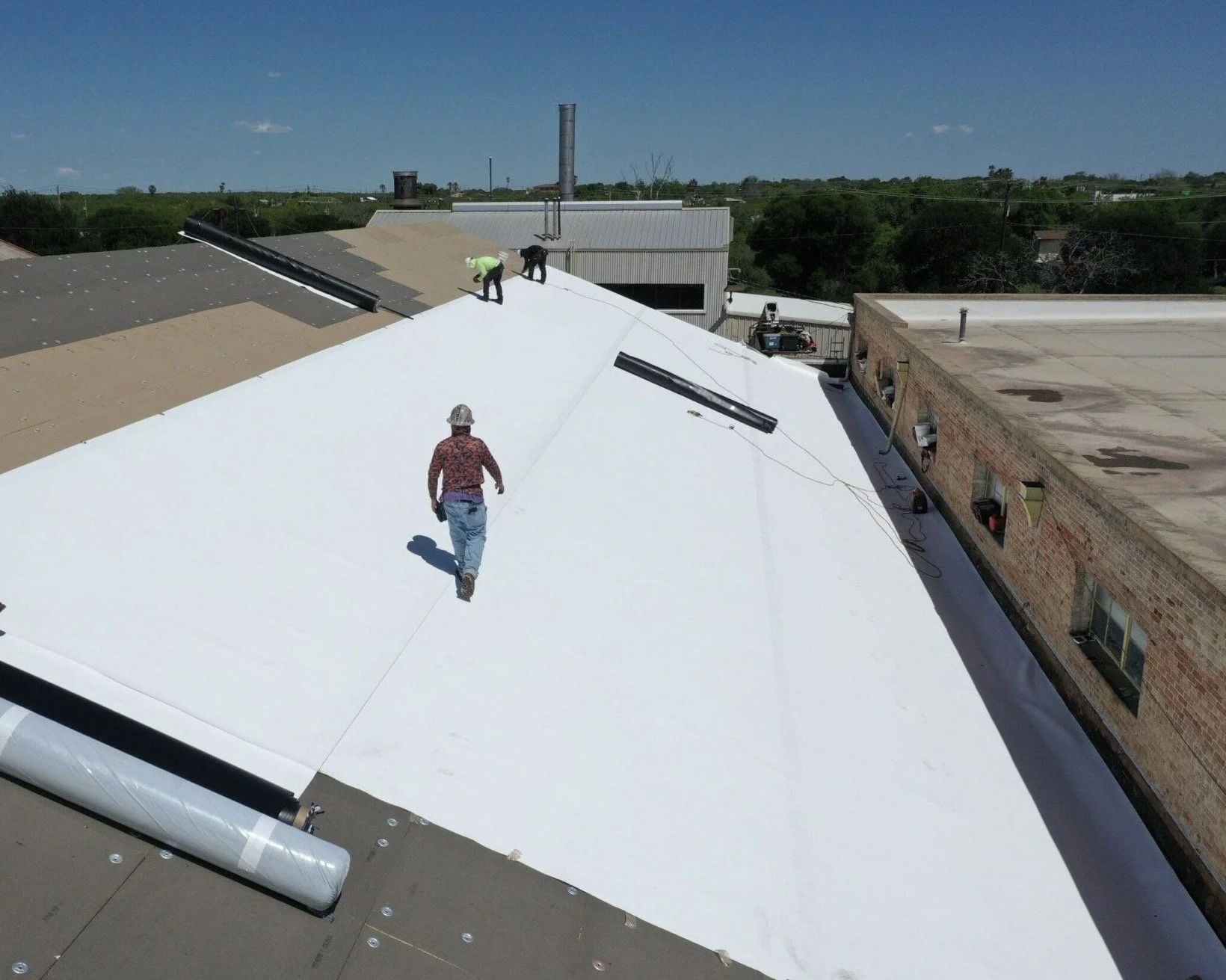 Workers installing a reflective white roofing membrane on a building roof during the daytime.