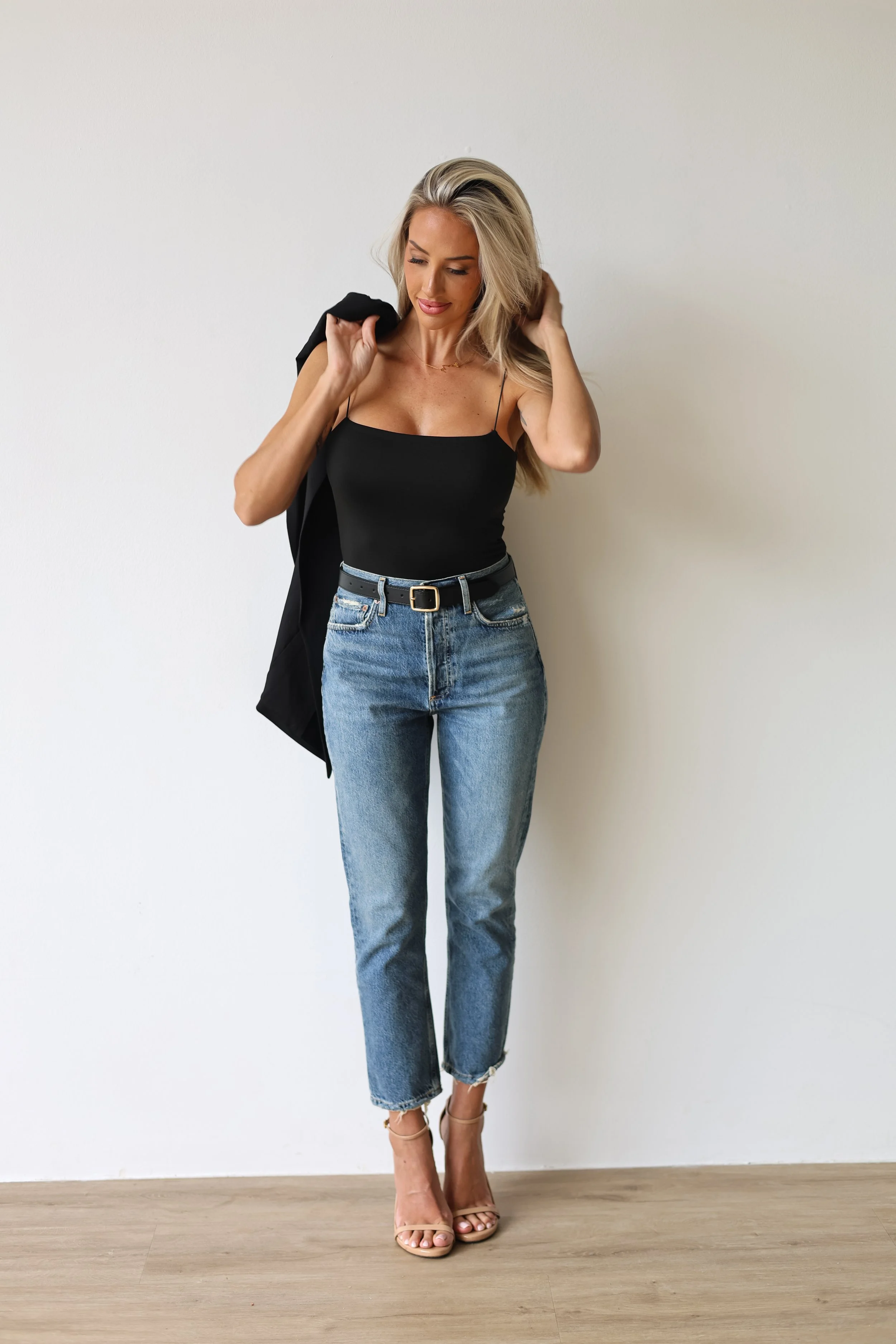 A woman with long blonde hair wearing a black spaghetti strap top, high-waisted blue jeans, tan high-heeled sandals, standing against a plain white wall, adjusting her hair with a black blazer draped over her shoulder.