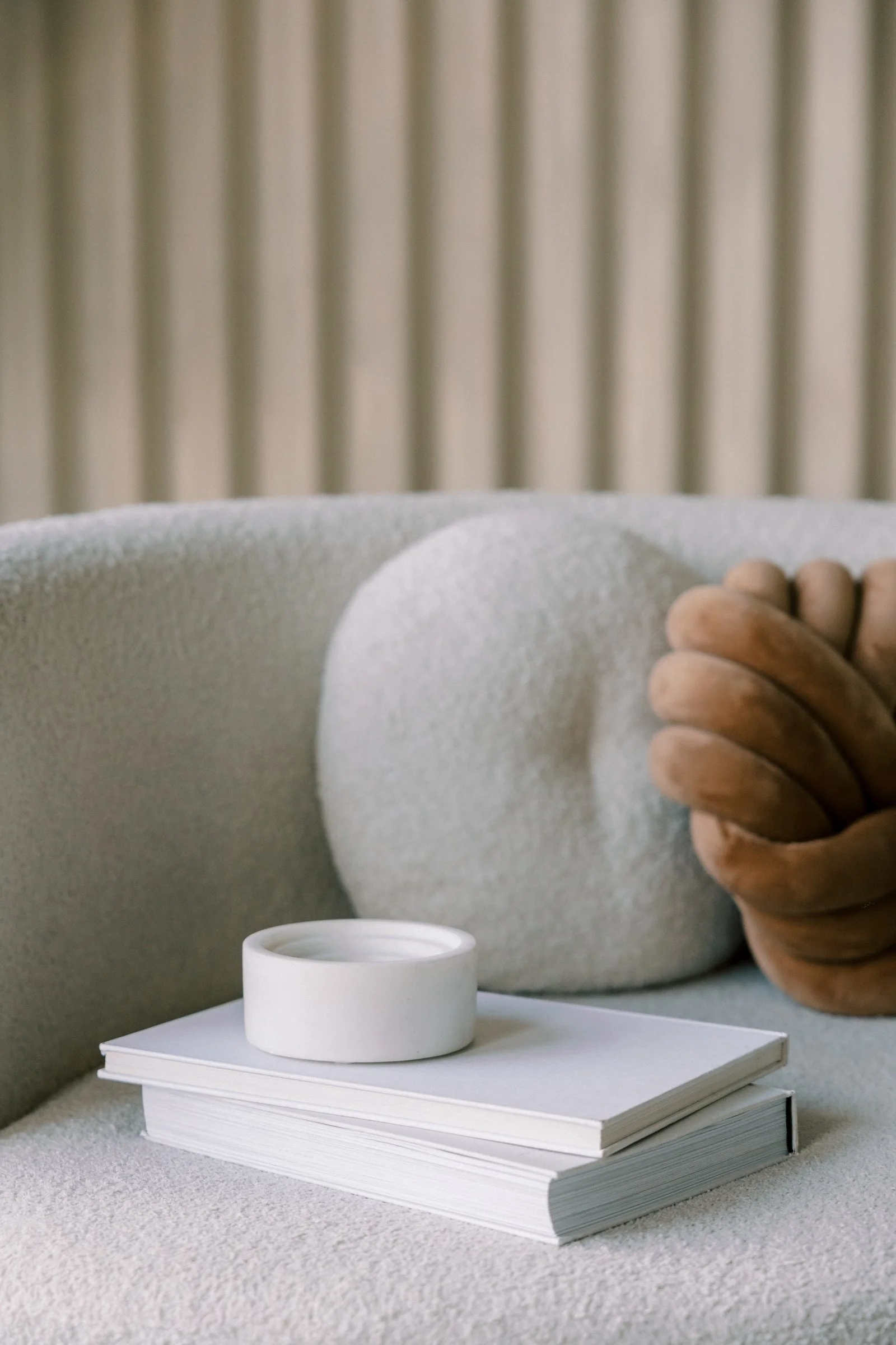A white ceramic bowl placed on top of a stack of white books on a white table in a room with a cream-colored sofa and a pillow, with vertical beige curtains in the background.