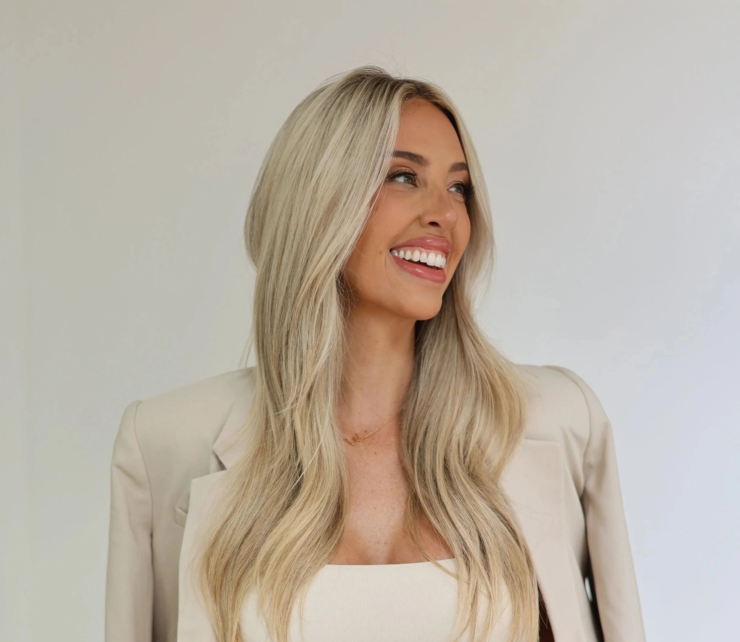 A woman with long blonde hair smiling, wearing a light-colored blazer and a white top, against a plain light background.