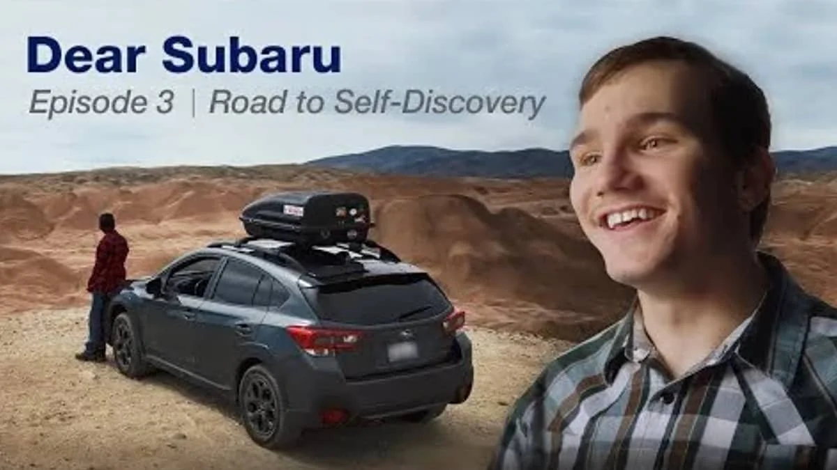 A smiling man in a plaid shirt standing in front of a black Subaru car with a roof cargo box, parked on a desert landscape with distant mountains. A second person in a red jacket stands beside the car, looking away.