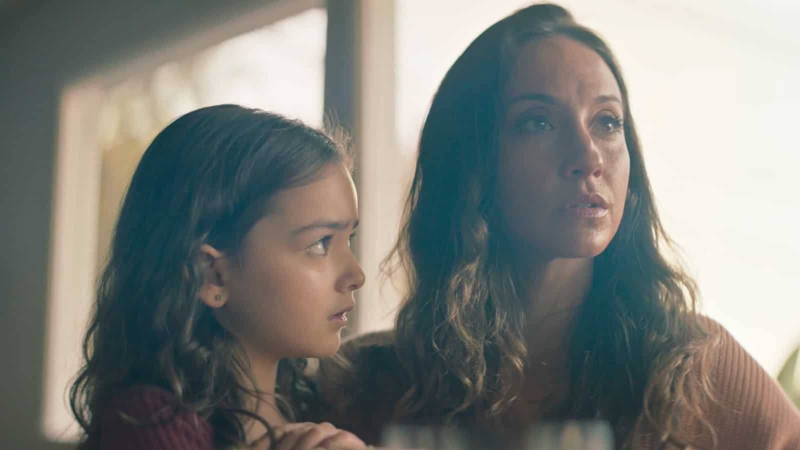 A woman and a young girl are sitting together indoors, with the woman speaking and the girl listening attentively. Both appear serious and engaged in the moment.