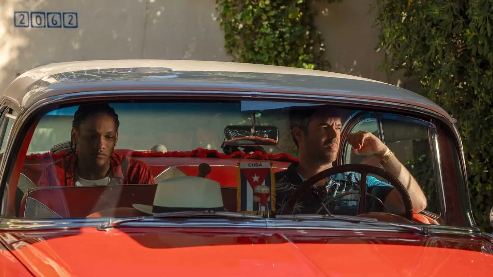Two sitting in a vintage red-colored car. The man is driving and seems to be somber, while the other is sitting as a passenger with a serious expression. There is a small Cuban flag hanging from the rearview mirror.