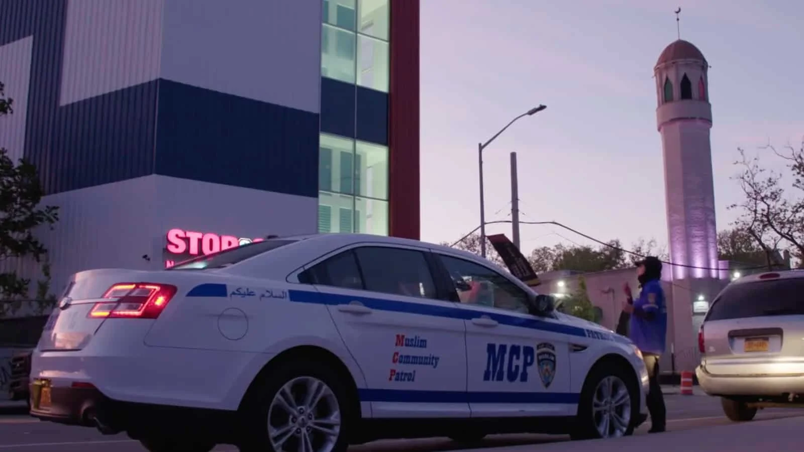 A police car with lettering Muslim Community Patrol parked on the street at dusk in front of a building and a tall tower with a dome, with a person standing nearby.