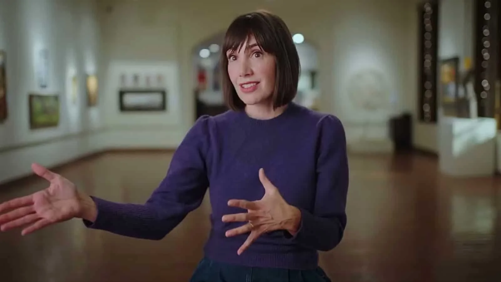 A woman with short dark hair wearing a purple sweater, gesturing with her hands while speaking in an art gallery or museum.