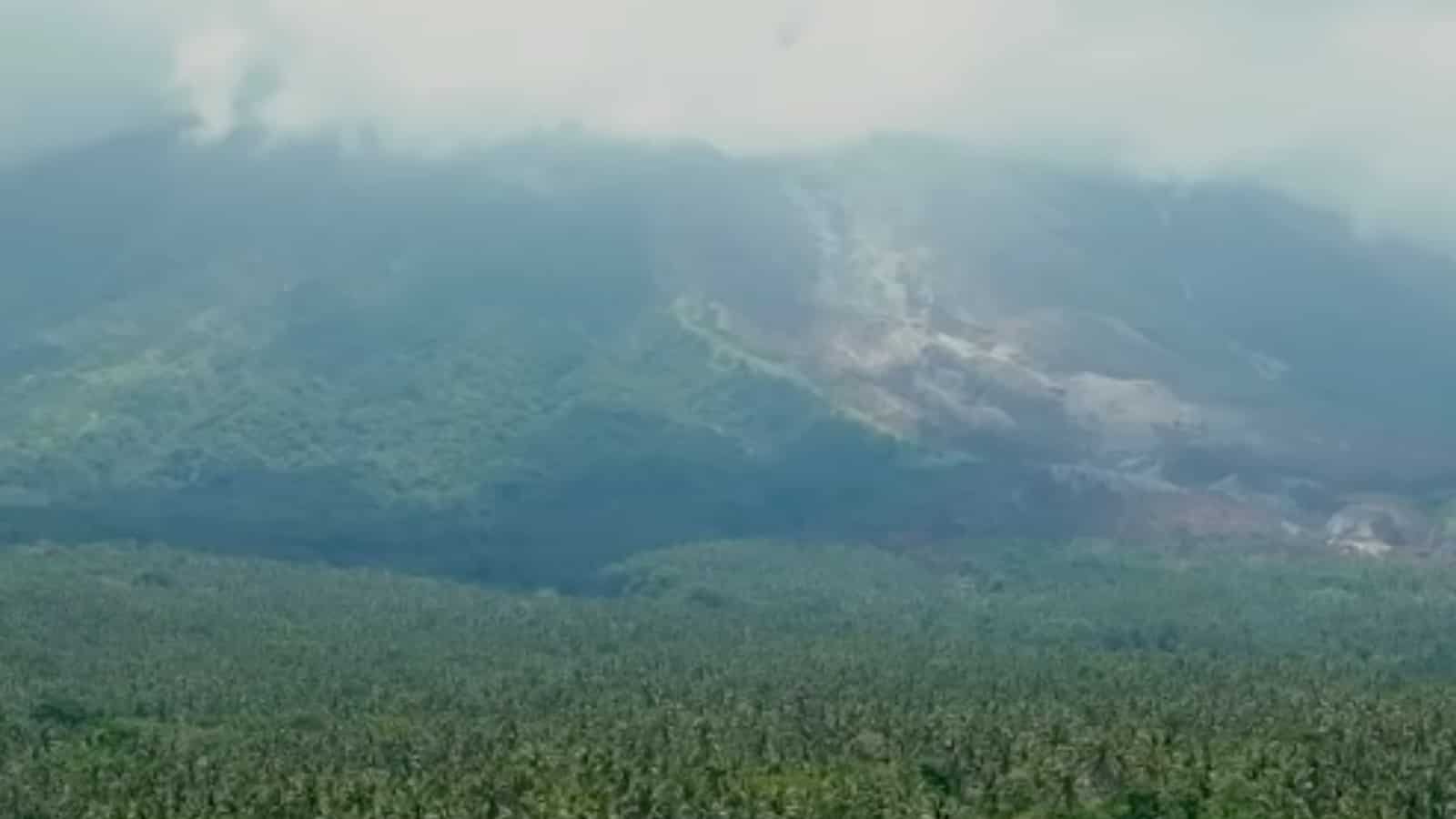 A mountainside with patches of smoke and visible signs of a wildfire, surrounded by a forested landscape under a cloudy sky.