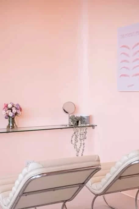 A pink-colored room with a glass shelf on the wall holding a small mirror. There is a bouquet of pink and purple flowers in a vase on the shelf. Part of a poster with eyebrow illustrations is visible on the wall. Two white chairs with ornate backs are in the foreground.