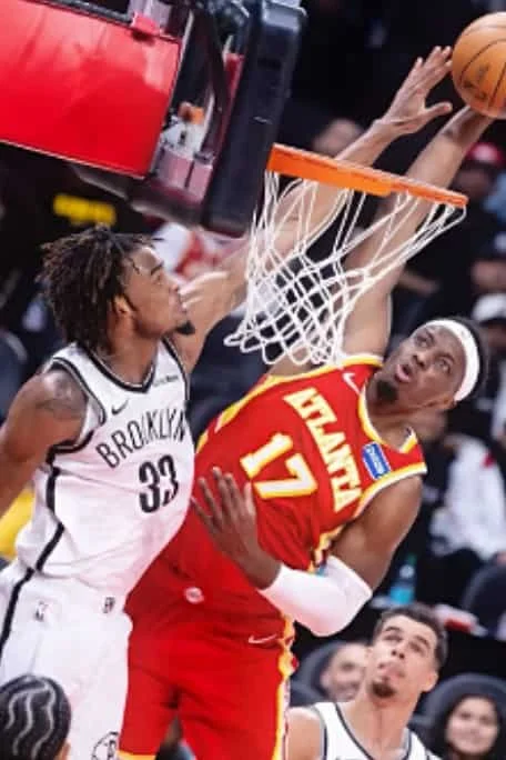 A basketball player in a red Atlanta Hawks uniform is attempting a shot while another player in a Brooklyn Nets uniform blocks it during a game.