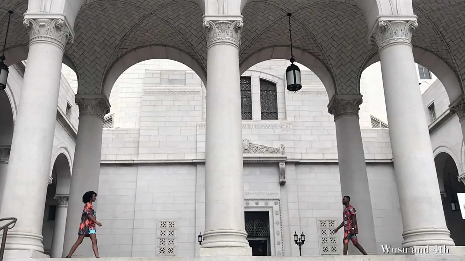 Two women walking past large white columns of a classical-style building with arches and ornate details in an urban setting.