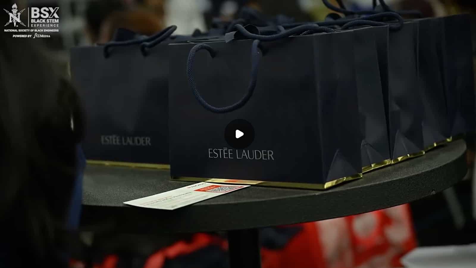 Black gift bags on a table, with the Estée Lauder logo, at an event for the Black STEM Experience, organized by the National Society of Black Engineers, with a white event ticket in front of the bags.