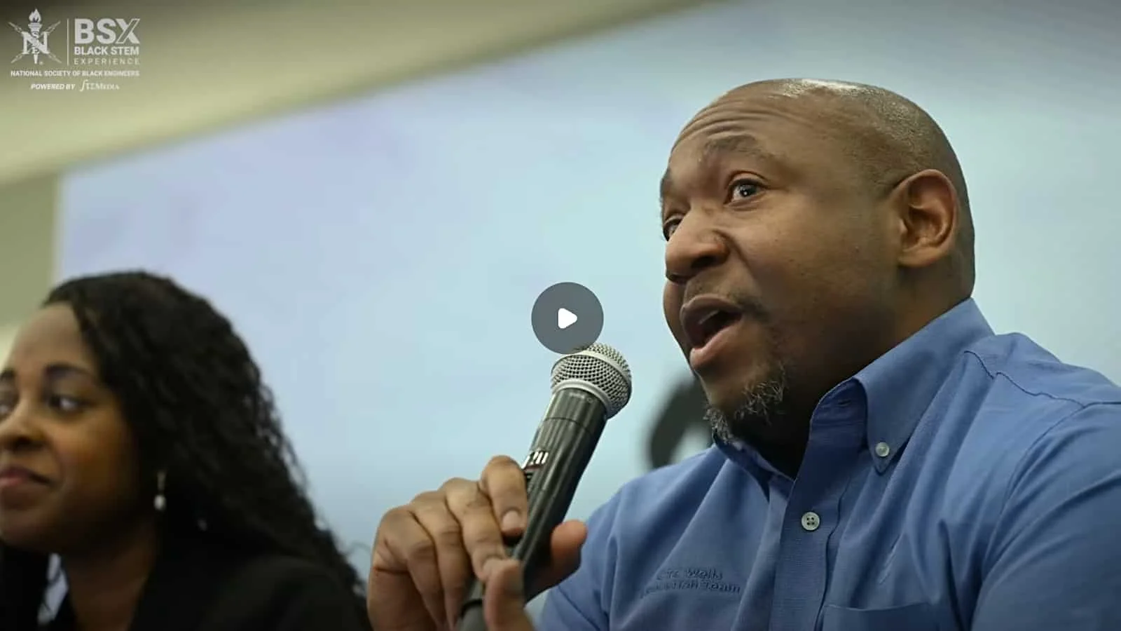 A man speaking into a microphone during a panel discussion, with a woman sitting beside him. The background includes a logo for Black STEM Experience.