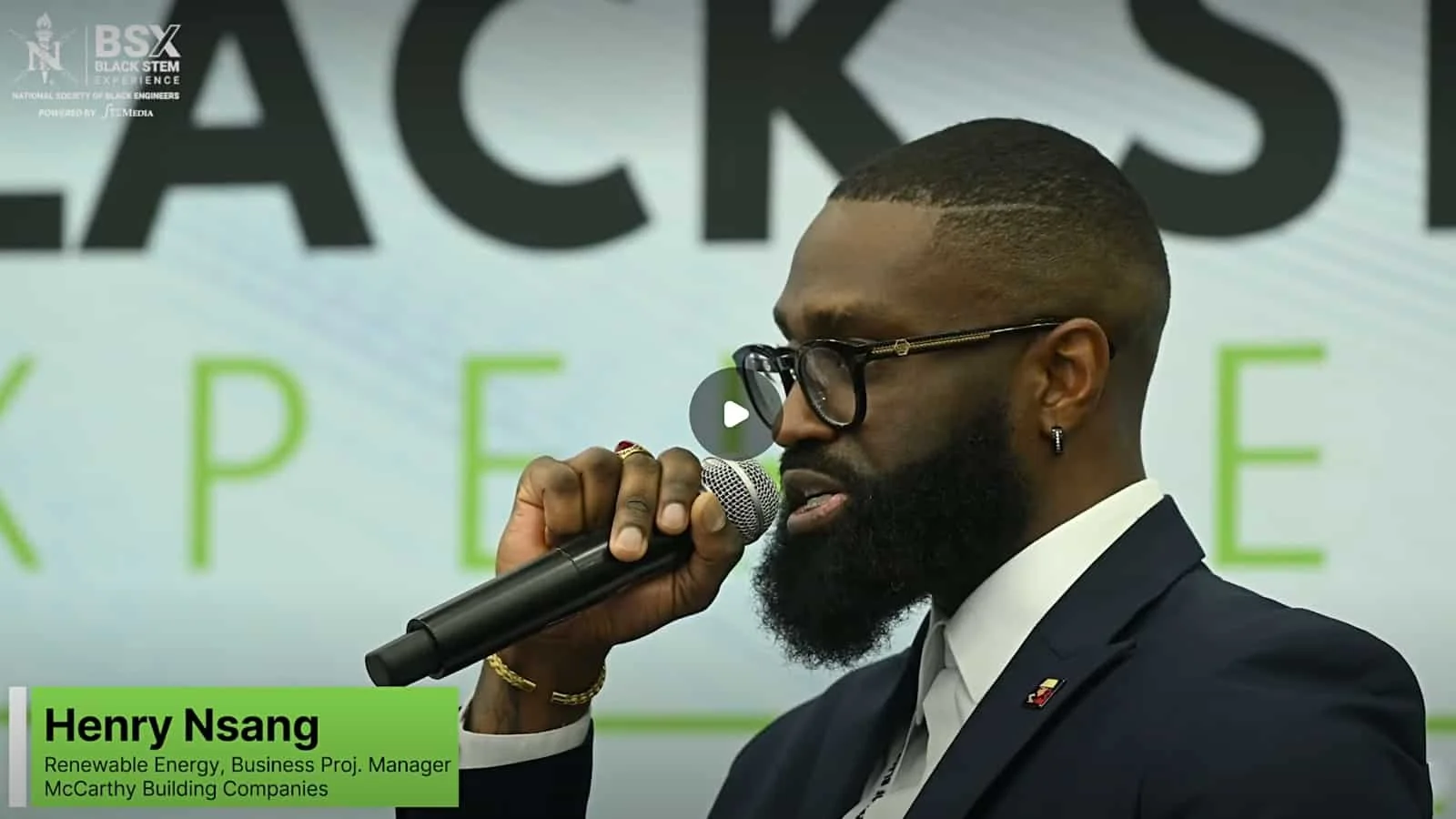 A man with a beard, glasses, and earrings holding a microphone and speaking at an event. He is dressed in a dark suit and white shirt. The background shows large text with the words 'BLACK STEM' and 'Experience'.