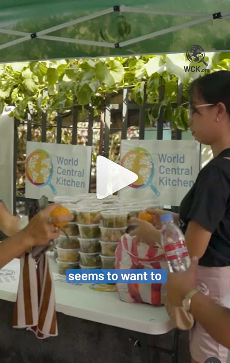 People at a food distribution table with containers of food and bottled water, with sign reading 'World Central Kitchen' in the background.