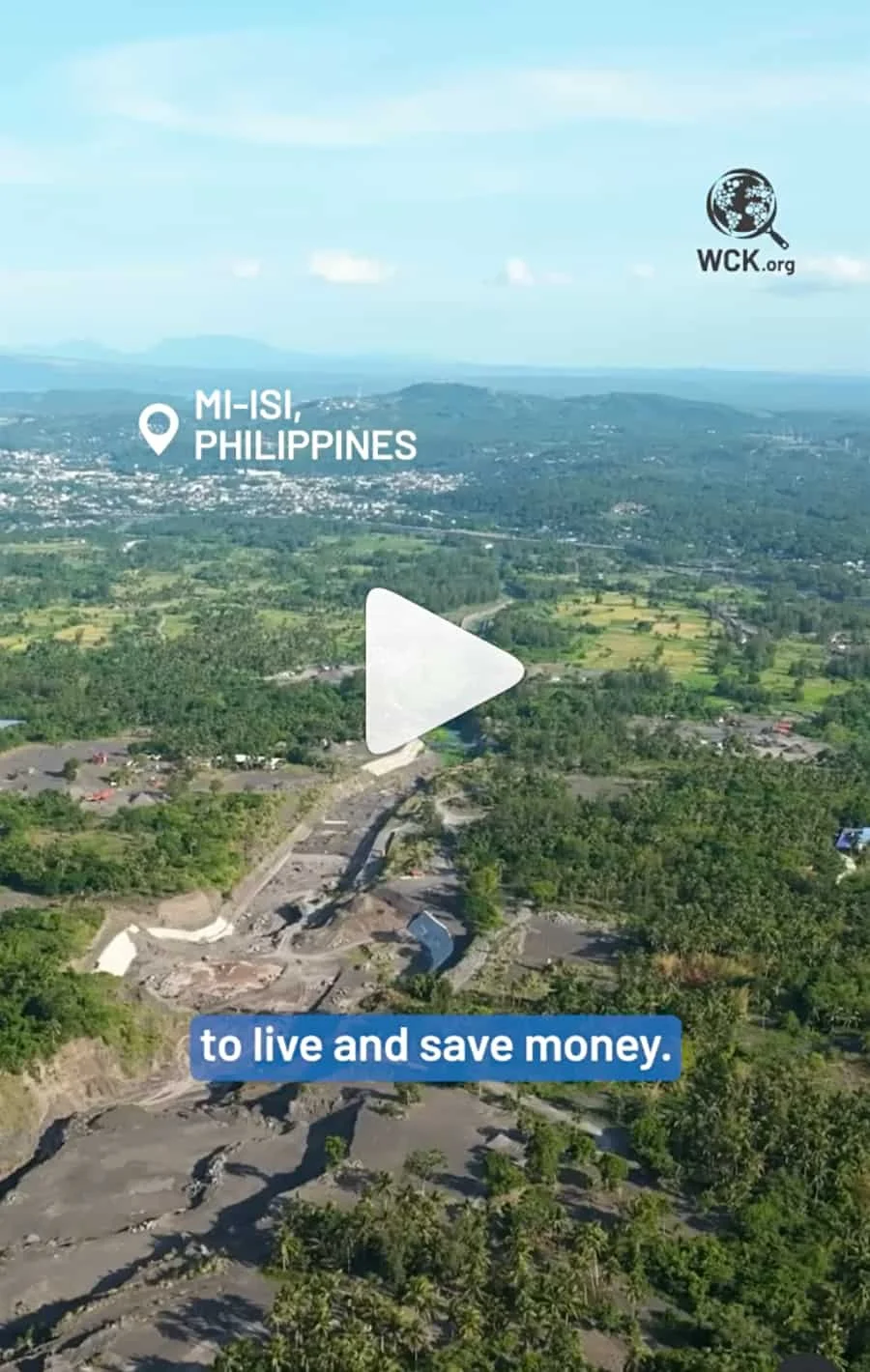 Aerial view of a lush, green landscape in M-Isi, Philippines, with a mix of trees, fields, and small buildings, under a bright blue sky with some clouds.