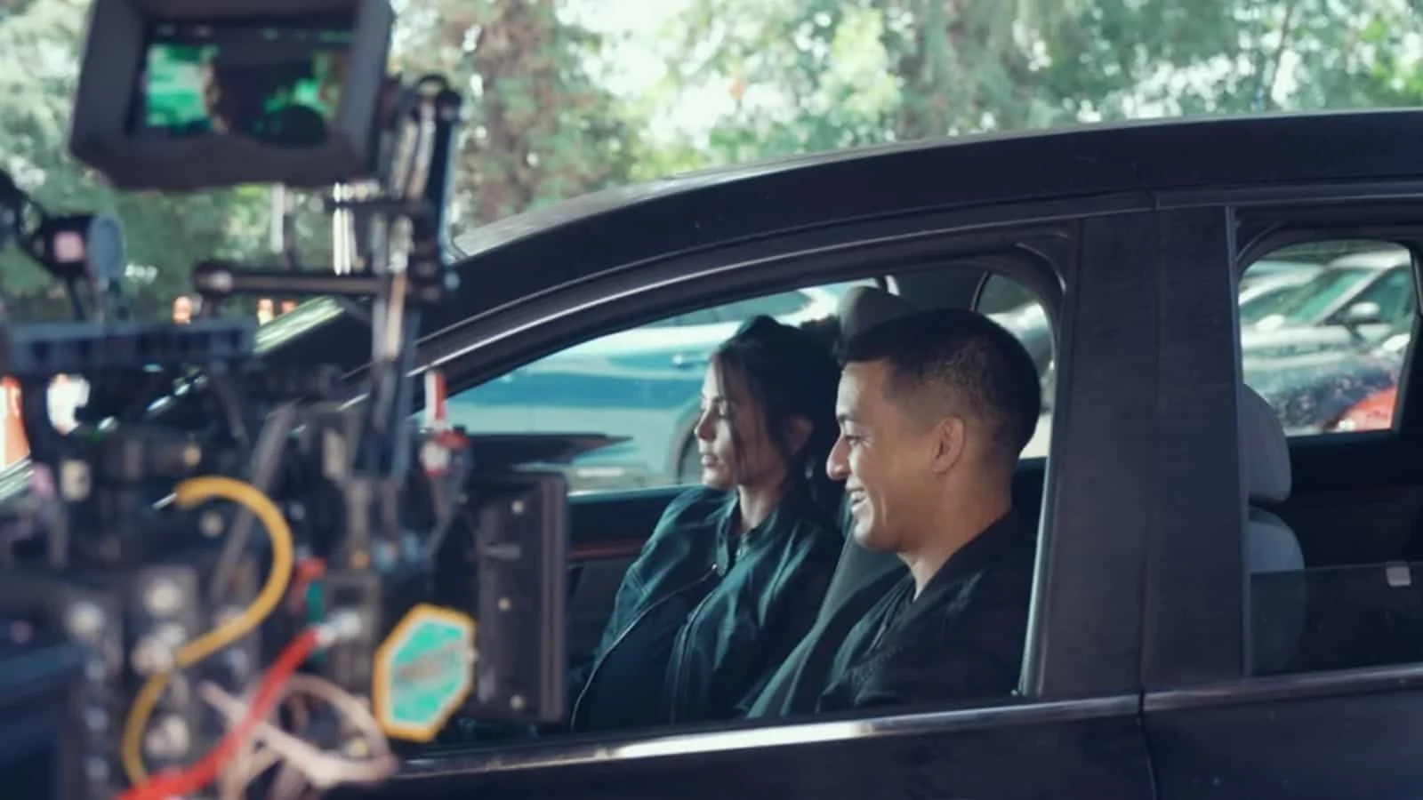 Two people smiling while sitting inside a car, with a camera setup visible outside the car recording them.