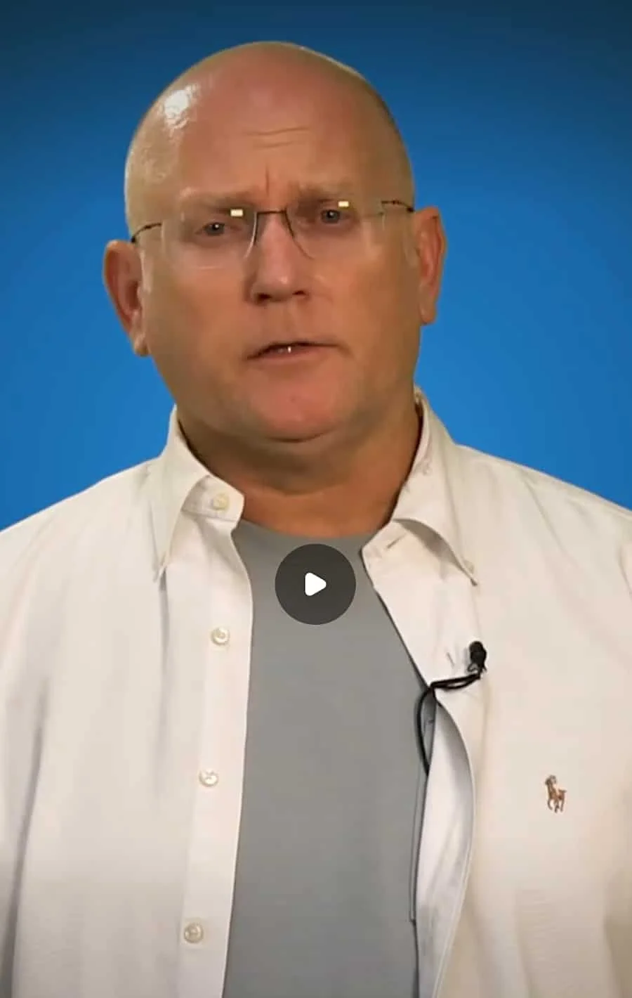 A middle-aged man with glasses and a bald head, wearing a white button-up shirt over a gray t-shirt, speaking against a blue background.