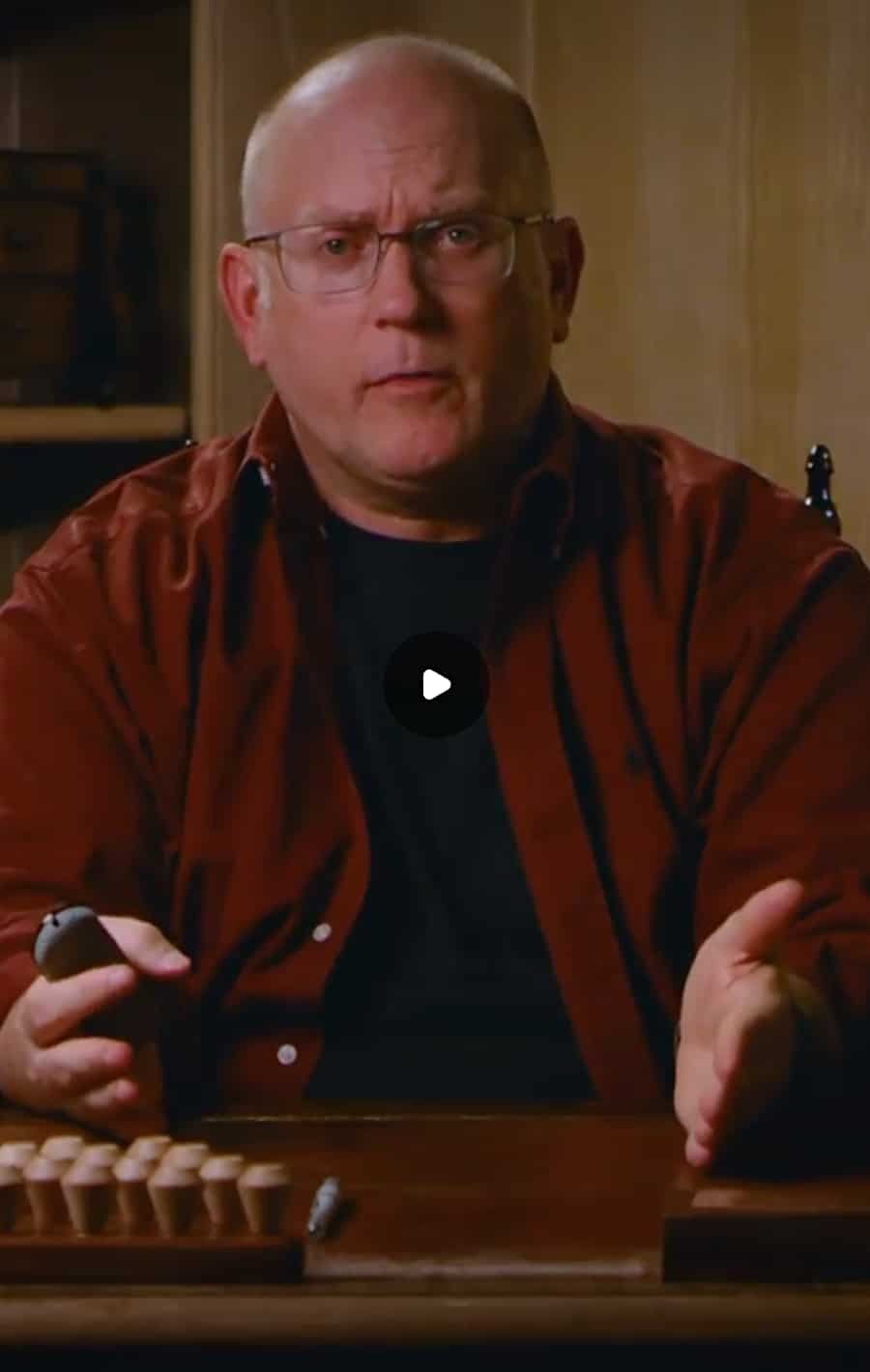 A man with glasses and a bald head wearing a dark red shirt, sitting at a table with a game board, speaking with a serious expression.
