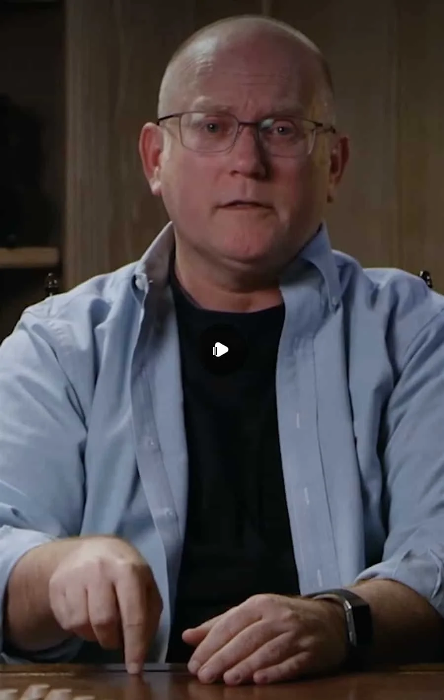 A middle-aged man with glasses, wearing a black t-shirt and a light blue button-up shirt, sitting at a wooden table in a room with wood paneling background.