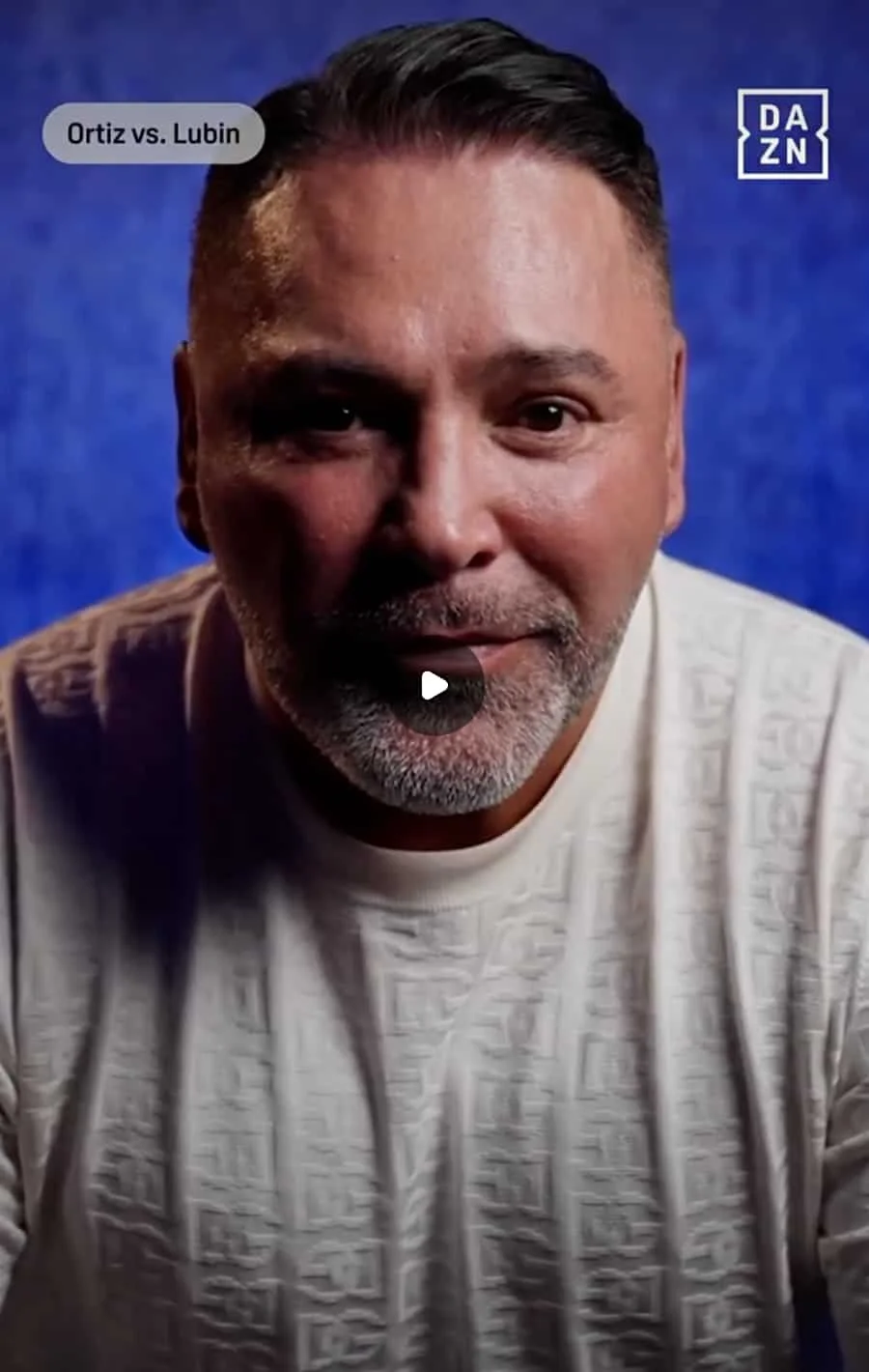 Close-up of Oscar De La Hoya with short dark hair, a beard, and a mustache, wearing a light-colored shirt, against a blue background. Text on the image reads "Ortiz vs. Lubin" and the DAZN logo is visible.
