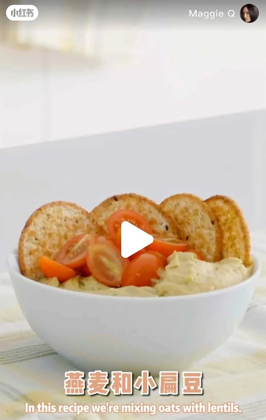 A white bowl filled with a dish that includes hummus, cherry tomatoes, and topped with round toasted crackers.