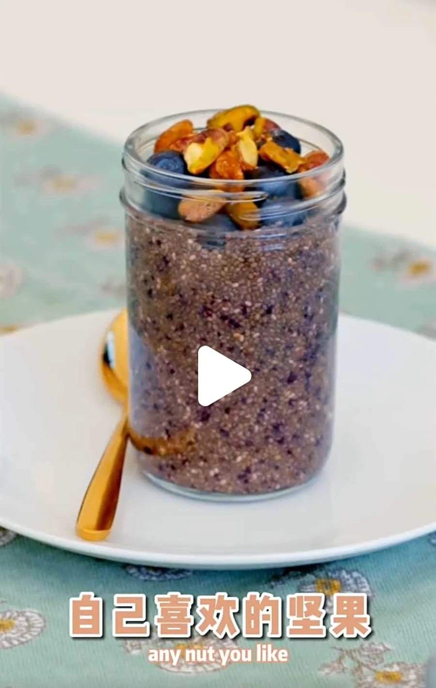 A glass jar filled with a mixture of crushed nuts and seeds, topped with mixed nuts and berries, placed on a white plate with a golden spoon next to it, on a light-colored tablecloth with floral pattern.