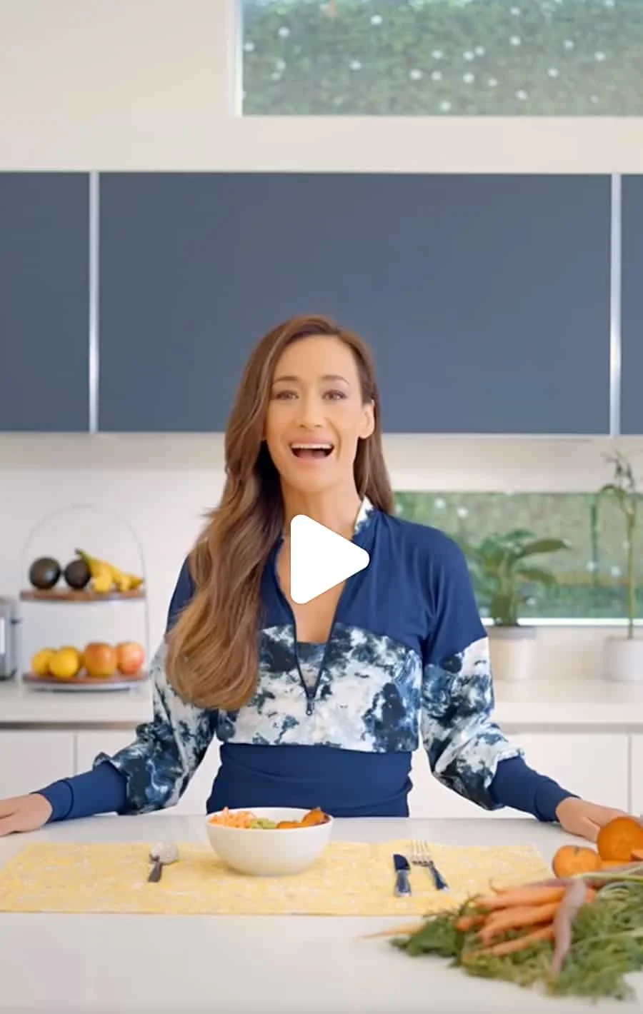 A woman standing behind a kitchen counter, smiling, with a bowl of food, cut vegetables, and fresh produce around her.