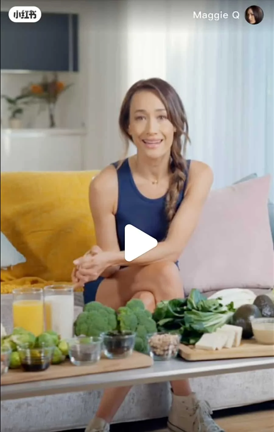 A woman sitting on couch with a yellow blanket and pink pillow, with a variety of groceries on a table in front of her, including vegetables, fruits, and beverages.