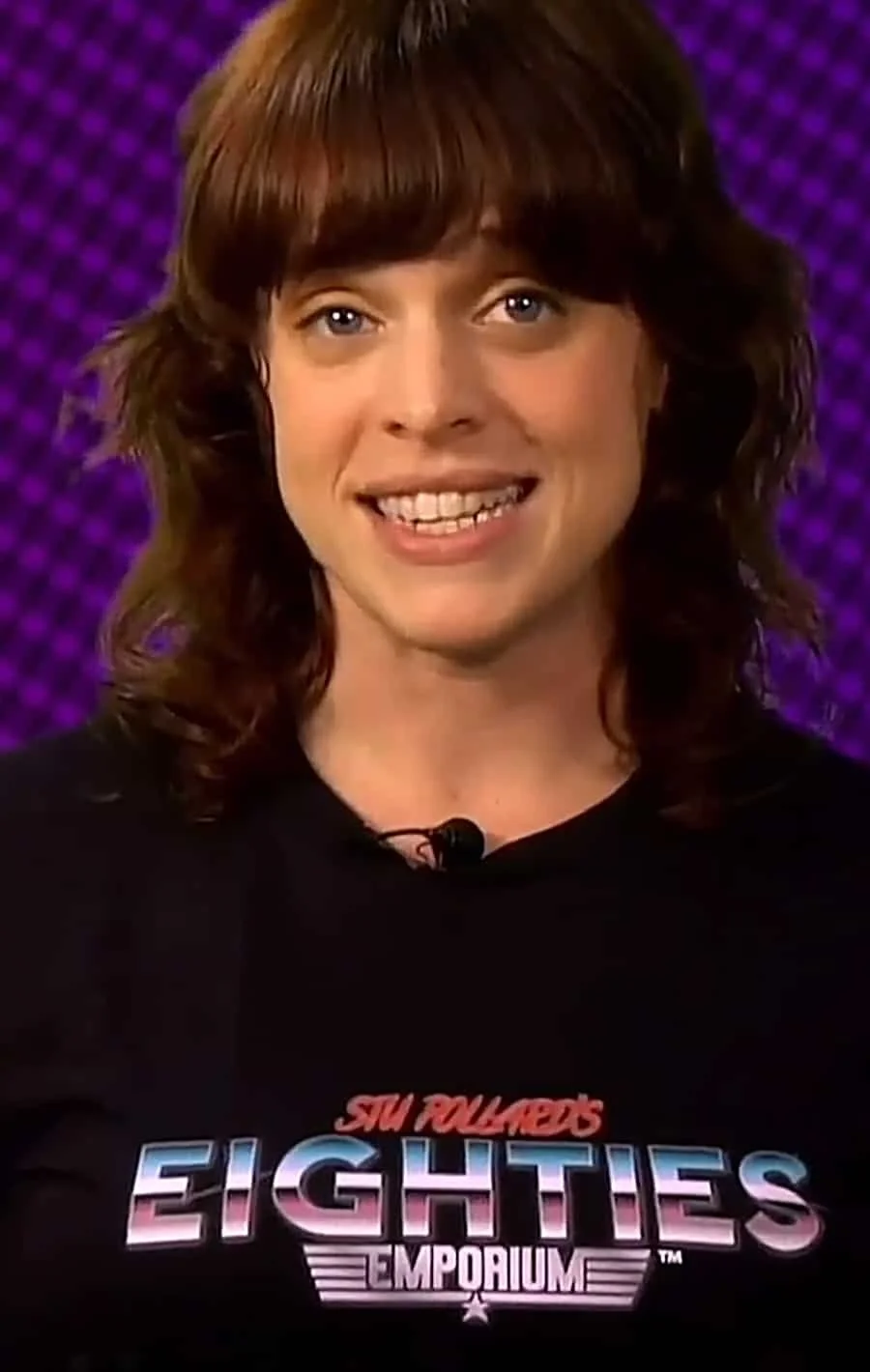 A woman with shoulder-length brown hair, smiling while wearing a black T-shirt with a logo that says "EIGHTIES EMPORIUM" in colorful text, in front of a purple background.