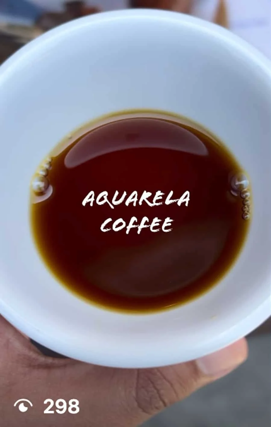 Close-up of a white cup filled with dark coffee, with the words 'Aquarela Coffee' written across the surface, and a hand holding the cup visible at the bottom of the image.