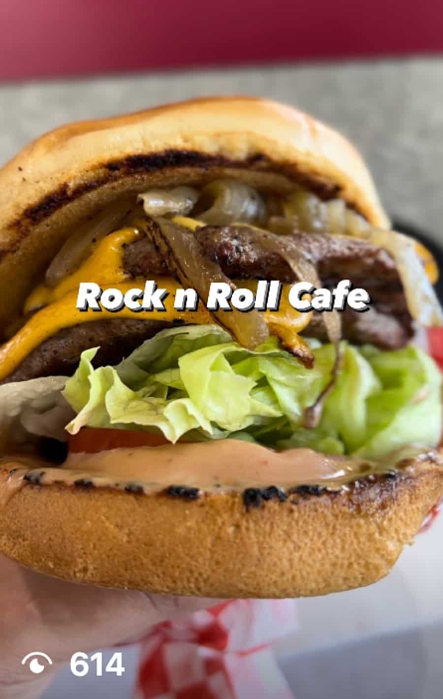 Close-up of a cheeseburger with lettuce, tomato, grilled onions, melted cheddar cheese, and grilled beef patties in a toasted bun, branded with Rock n Roll Cafe.