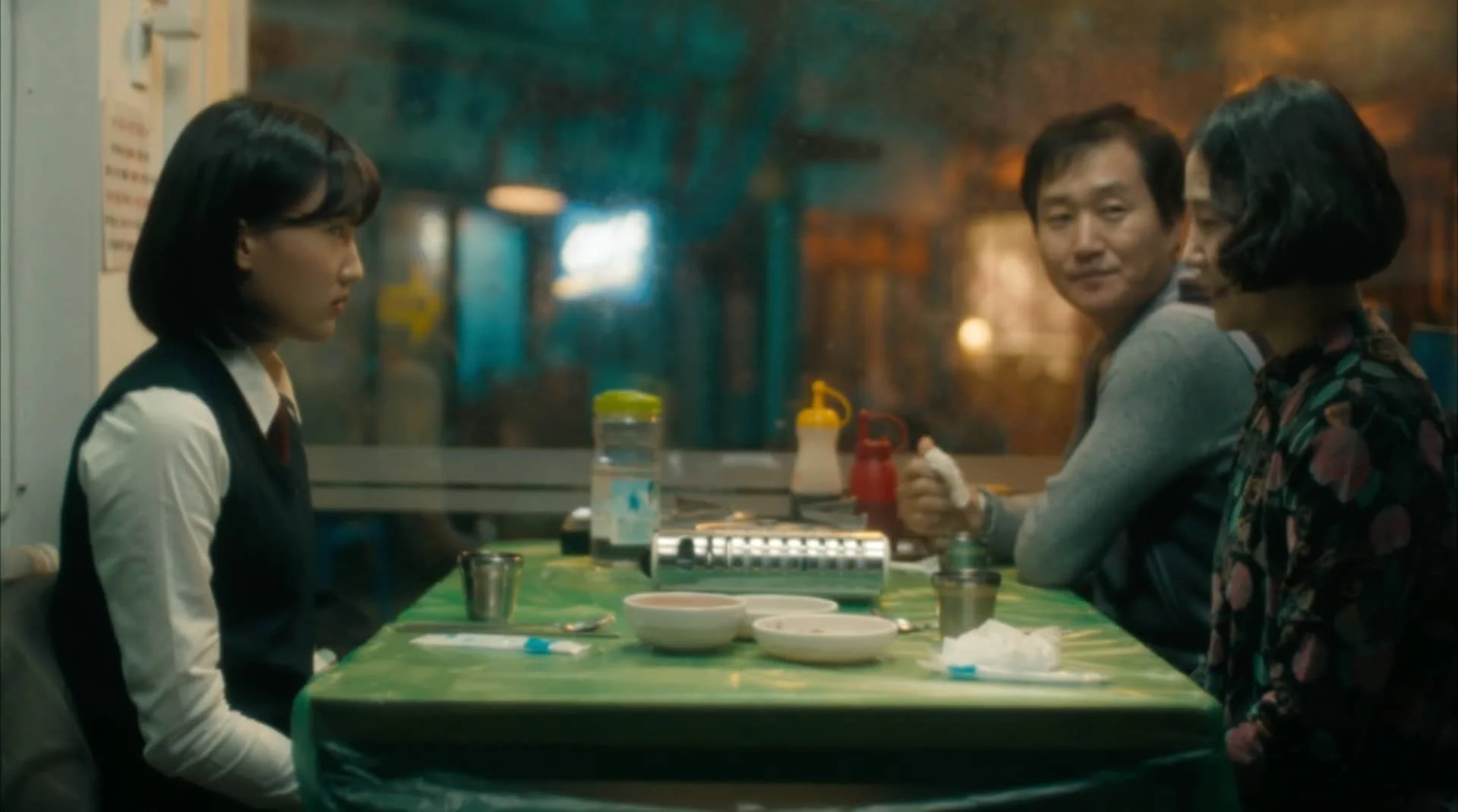 Three people sitting at a green table in a restaurant or diner, with one woman on the left facing two people on the right, a man and a woman, with condiment bottles and bowls on the table, and a window or glass separating the scene from the outside.