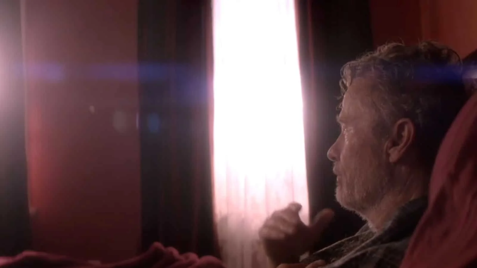 A man with gray hair and beard lying on his side on a bed, looking at a bright light coming through a curtain.