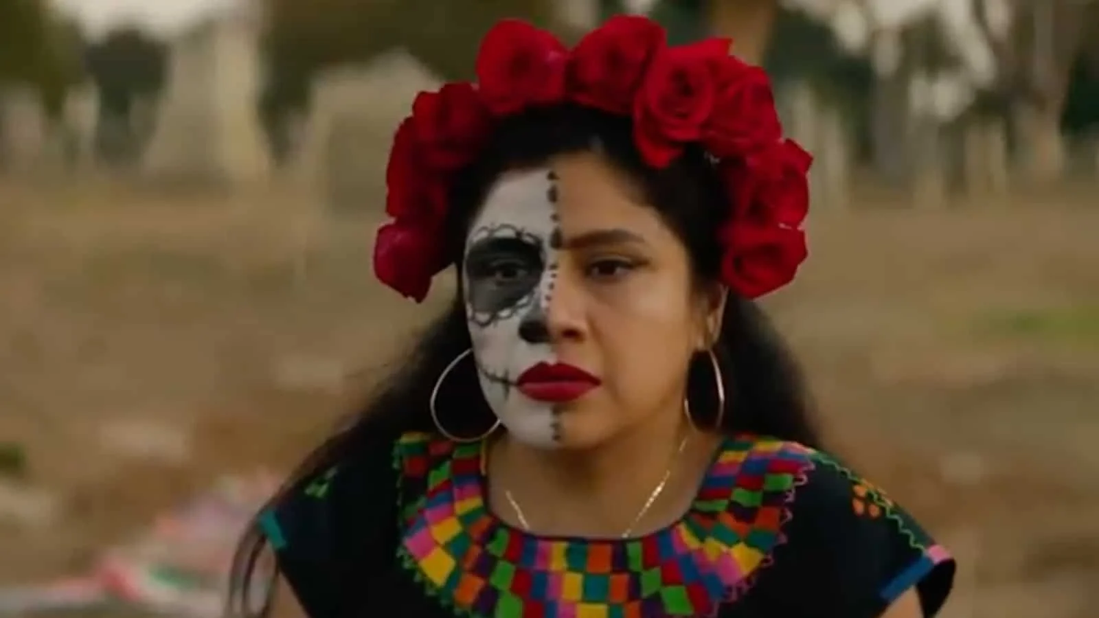 A woman with half her face painted in a skull design, wearing red roses on her head, jewelry, and a colorful embroidered top.