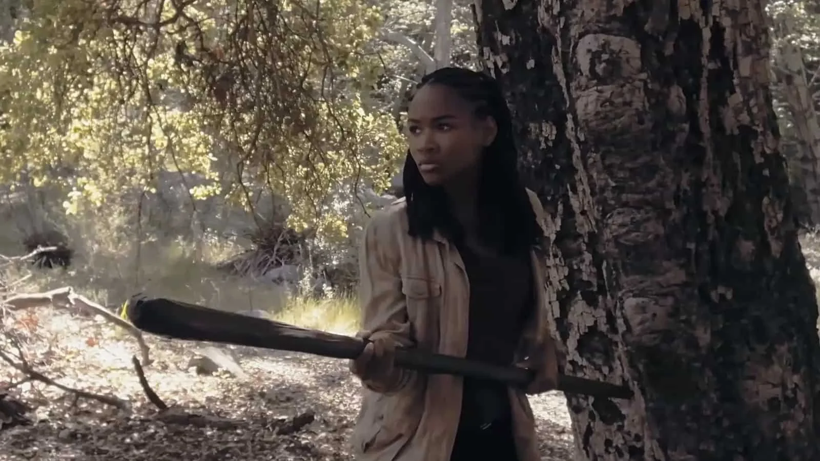A woman with braided hair holding a stick or bat, looks to be confronting an unseen character in a wooded area