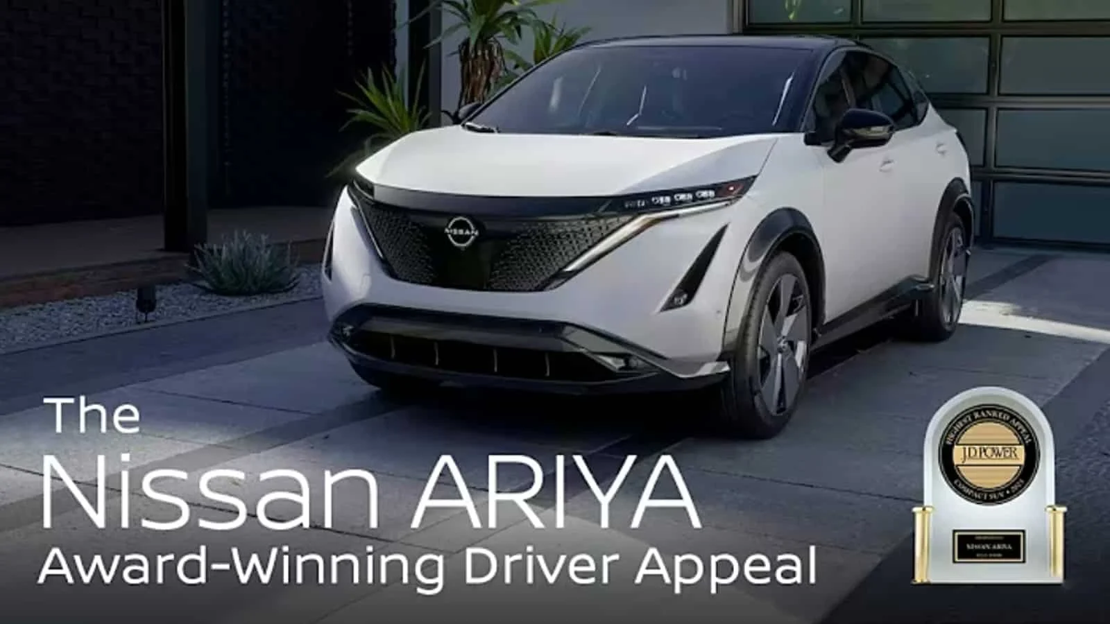 A white Nissan Ariya electric vehicle parked in a driveway next to a modern house with a large window and a plant. The overlay text reads 'The Nissan ARIYA Award-Winning Driver Appeal' with an award emblem on the bottom right.