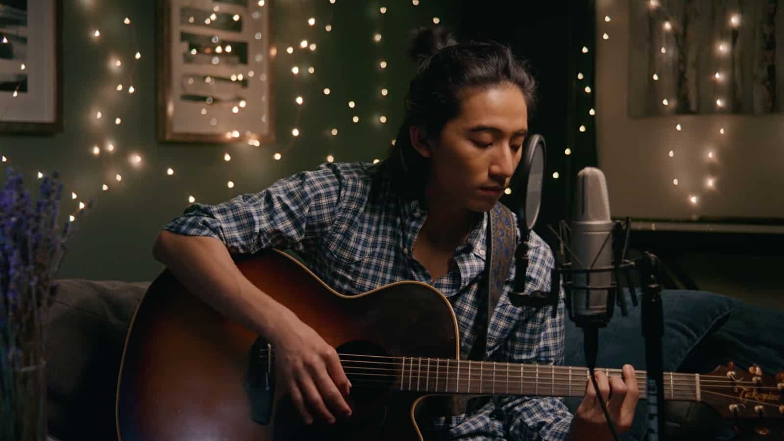A man with long dark hair playing an acoustic guitar in a dimly lit room decorated with string lights and framed artwork, with a microphone positioned nearby.
