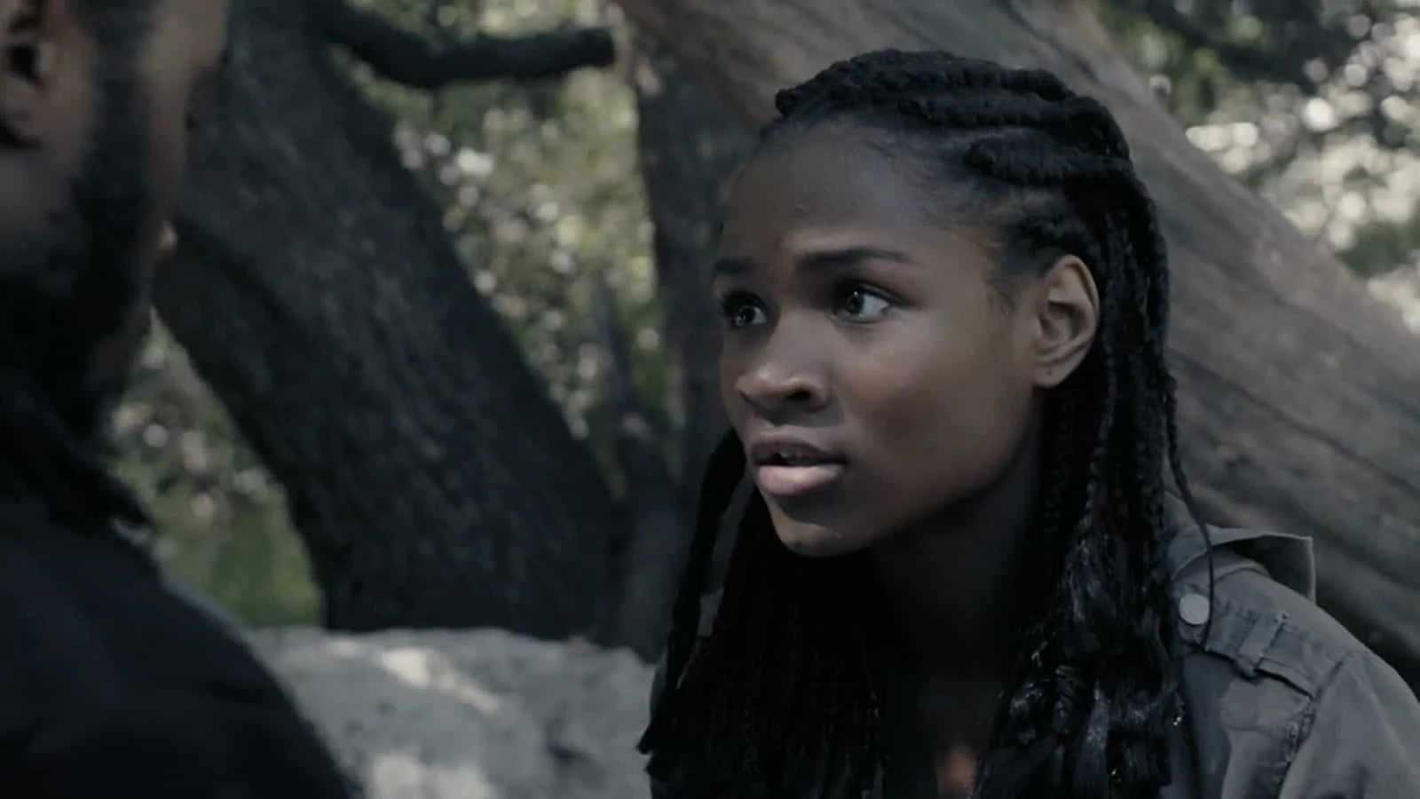A young woman with dark skin and braided hair looking surprised or concerned outdoors with trees and a fallen log in the background.
