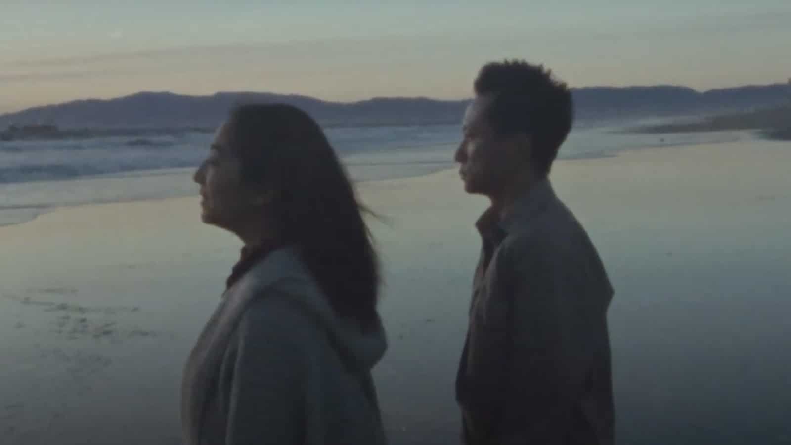 Two people, a woman and a man, walking along the beach at sunset or dusk, with ocean waves and distant hills in the background.