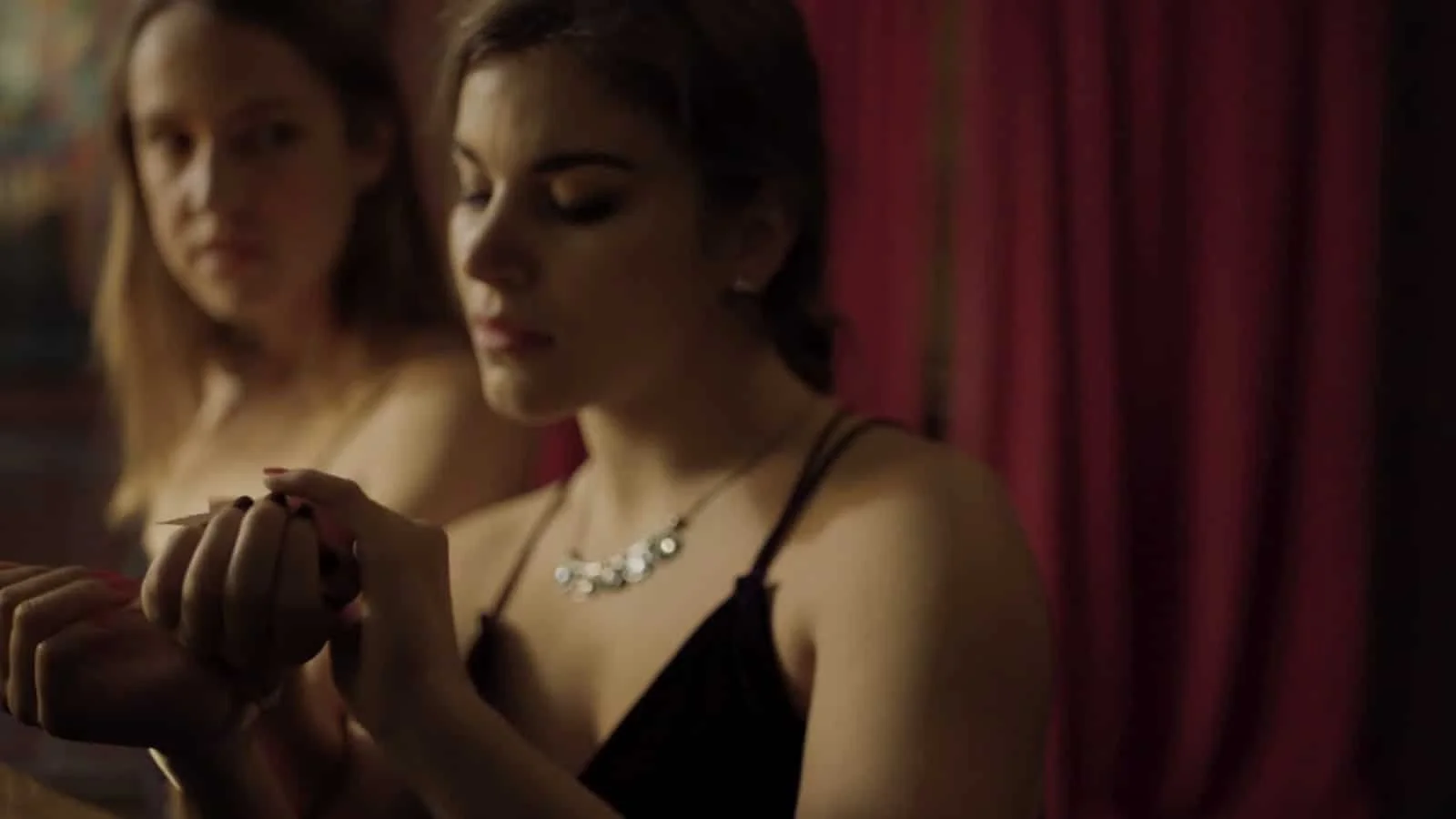 Two women are sitting close together in a dimly lit room with red curtains in the background. One woman is in focus, wearing a black dress with thin straps and a necklace. The other woman is slightly out of focus, looking at the first woman.