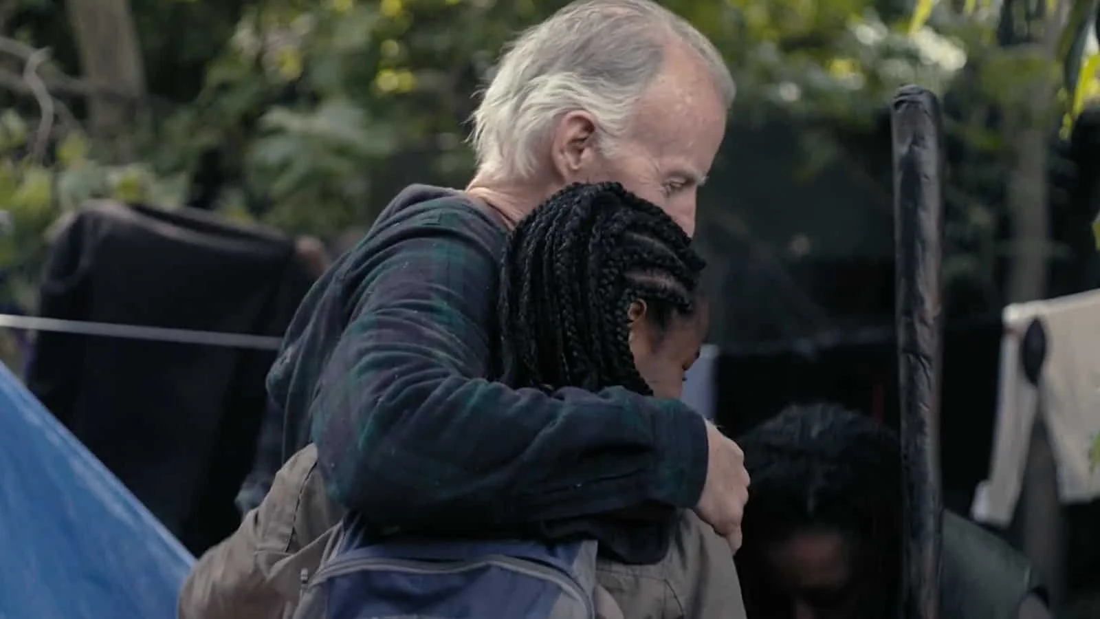 An elderly man with white hair hugging a young girl with braided hair in an outdoor setting with trees in the background.