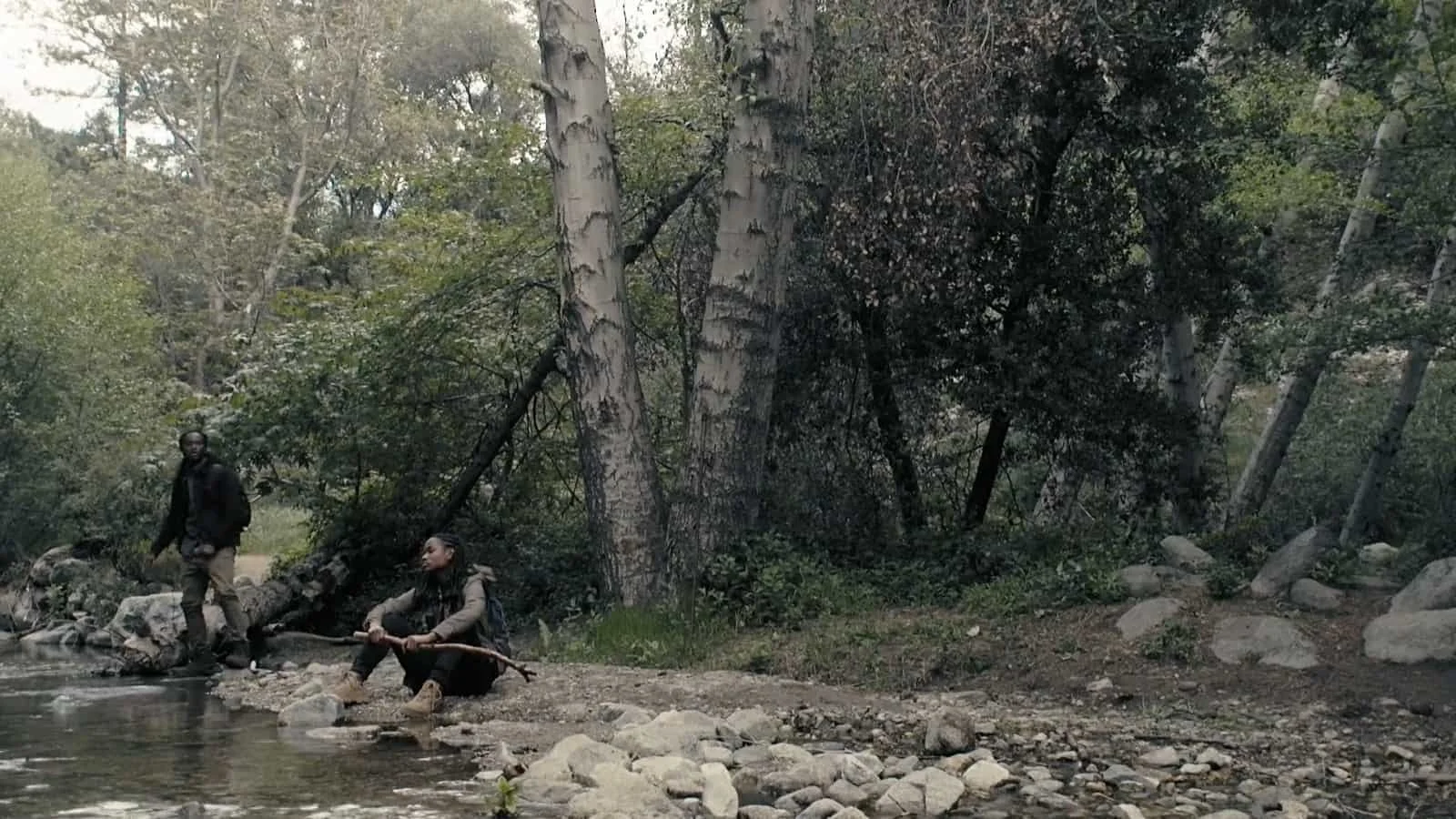 Two people are by a river in a forested area, one is standing on rocks next to the water and the other is sitting on the ground holding a stick, surrounded by tall trees and rocks.