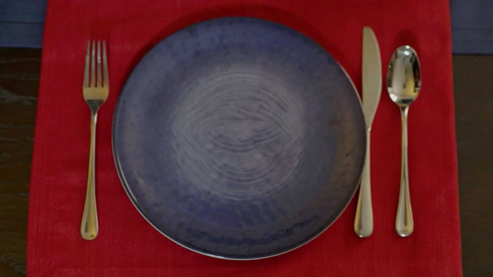 Empty blue ceramic plate on a red placemat with a fork on the left and a knife and spoon on the right.