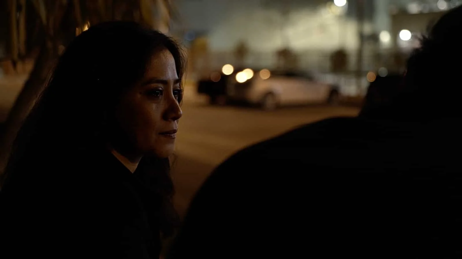 A woman with long dark hair sitting on a street at night, partially visible, with blurred parked cars and lights in the background.