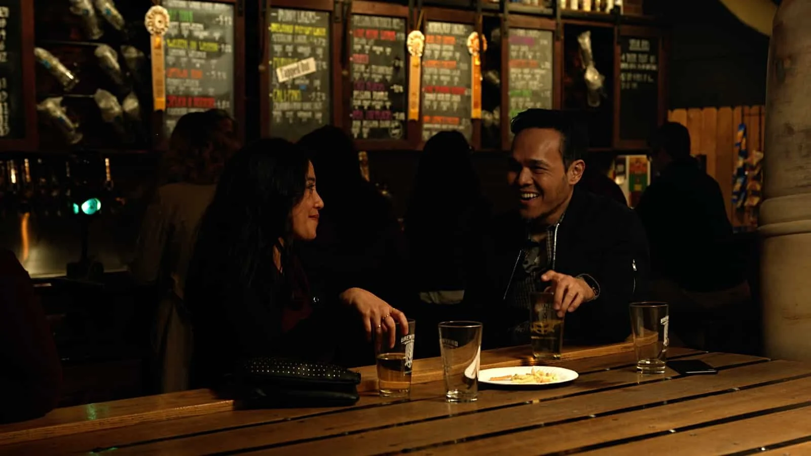 Two people, a woman and a man, sitting at a wooden table in a dimly lit bar, smiling and talking. There are four glasses and a plate on the table, with a menu board of drinks in the background.