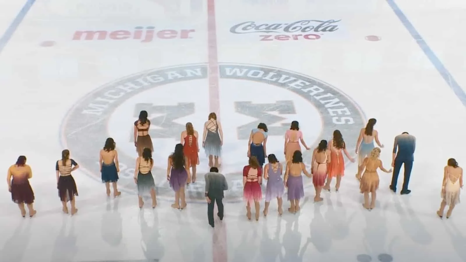 Group of people dressed in colorful costumes at the Michigan Wolverines ice hockey rink, standing around the center logo during a performance or event.
