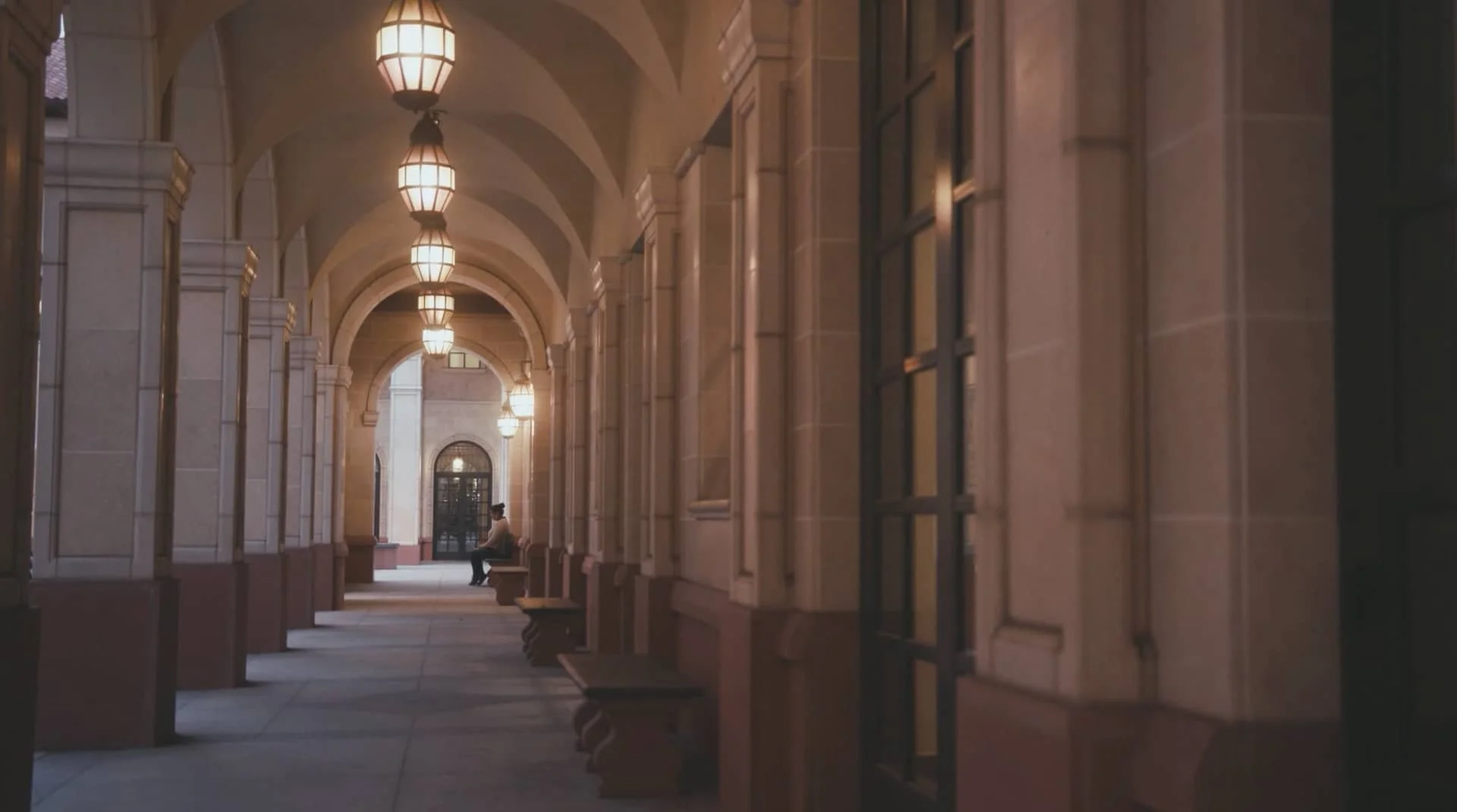 A person sitting on a bench in a covered walkway with arched ceilings and hanging lantern lights, with large windows and a door at the end.