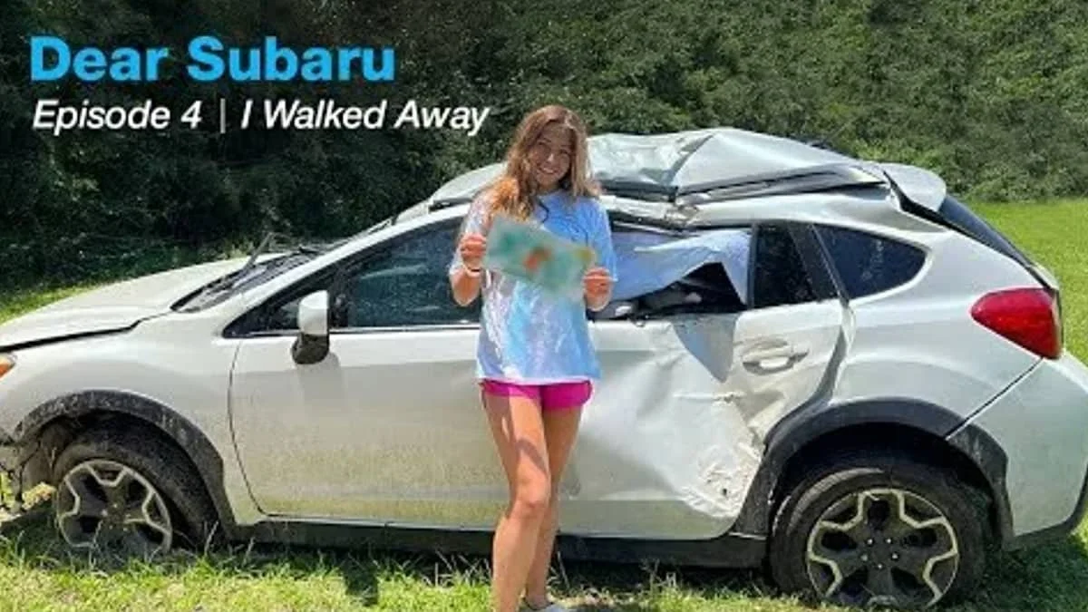 A young girl standing in front of a damaged Subaru car with a large dent and crushed roof, holding a map and smiling, outdoors in a grassy area.