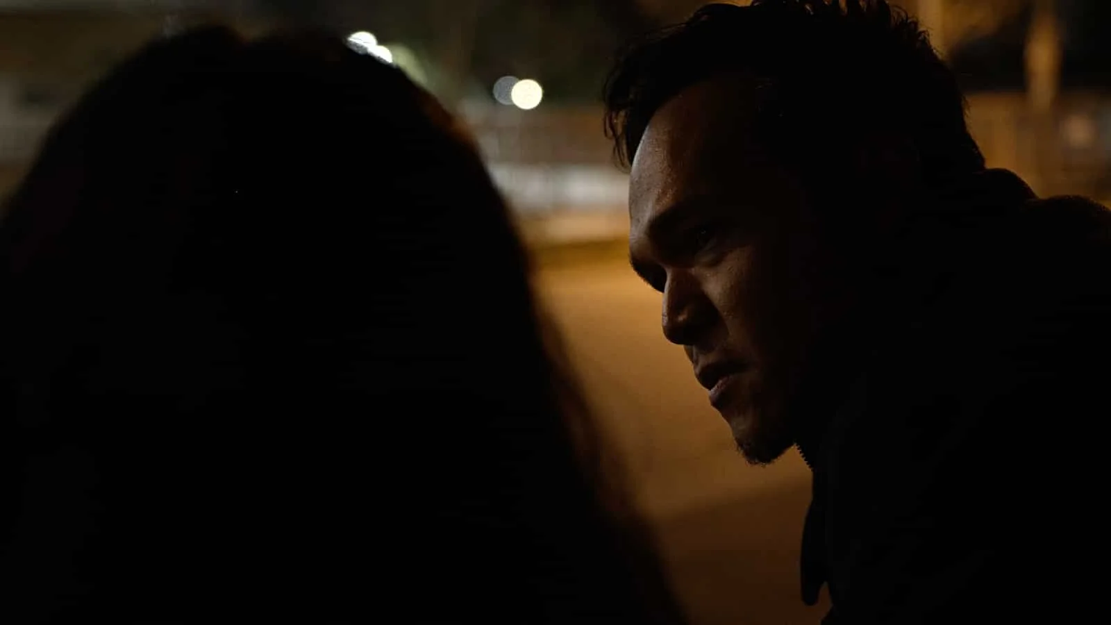 A man and a woman in a dark, outdoor setting in deep conversation, with the man intensely looking at the woman.